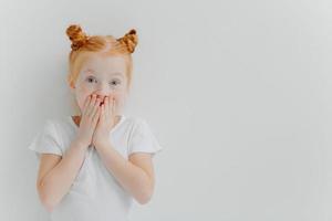 horizontaal schot van verrast roodharig meisje bedekt mond, kijkt naar spannende tekenfilm, heeft onverwachte reactie, draagt casual wit t-shirt, staat binnen, drukt verwondering uit. kinderen en emoties concept foto