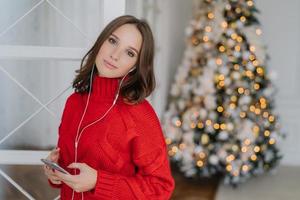 mooie jonge vrouwelijke model luistert aangename muziek in oortelefoons, houdt mobiel vast, gekleed in rode gebreide trui, poseert in woonkamer bij kerstboom met lege ruimte voor uw advertentie-inhoud foto