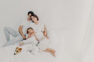 tevreden man, vrouw en hun kleine dochter poseren in ruime slaapkamer met favoriete huisdier, hebben plezier, omhelzen en uiten liefde voor elkaar, suppror in moeilijke situatie. familie tijd concept foto