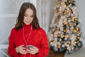 mensen en vakantie concept. geconcentreerde donkerharige jonge vrouw houdt mobiele telefoon in handen, leest nieuws op internet, gebruikt oortelefoons, staat binnen in de buurt van dennenboom, verbonden met snel internet foto