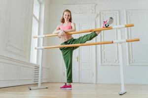 vrouwelijk model heeft aerobicsles, strekt de benen op balletbarre, draagt sportschoenen en sportkleding, leunt op de bar, glimlacht zachtjes, heeft rood haar, heeft repetitie. jonge ballerina traint in hal. ballet foto