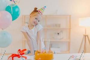 klein gembermeisje draagt feestmuts en wit t-shirt, staat in de buurt van feestelijke tafel met cake, blaast kaarsen en doet wensen tijdens haar verjaardag, poseert in witte kamer met opgeblazen ballonnen, glimlacht vreugdevol foto
