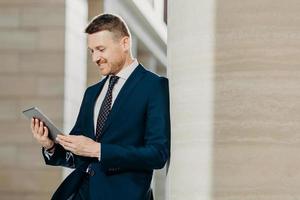 succesvolle mannelijke kantoormedewerker in zwart pak gebruikt moderne tabletcomputer voor afstandswerk, verbonden met draadloos internet, poseert in modern interieur, geconcentreerd in scherm. technologie concept foto