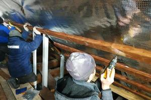 het team van de meesters installeert het dak van het huis en schroeft met name de schroeven in de planken voor het bevestigen van de foliefilm en minerale wol foto