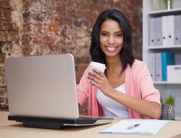 lachende vrouw met koffiekopje en laptop zit aan bureau