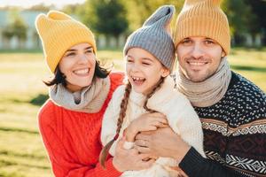 speels klein kind met staartjes draagt warme kleding, brengt vrije tijd door met lieve aanhankelijke ouders, heeft een gelukkige uitdrukking, voelt zich ontspannen. drie jonge gezinsleden genieten van aangename momenten foto