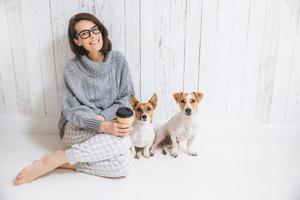 vrolijke blije brunette vrouw gekleed in warme winter gebreide kleding, zit in de kamer met haar honden, drinkt afhaalkoffie, poseert voor de camera, geniet van een goede huiselijke sfeer. vriendschappelijke relatie foto