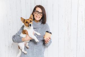 foto van tevreden brunette vrouw draagt haar favoriete hond, drinkt hete koffie uit een papieren beker, heeft een aangename warme glimlach op het gezicht, verheugt zich over de leuke tijd doorgebracht met huisdier, poseert tegen een witte omheiningsachtergrond