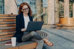 intelligente roodharige dame bladert buiten op laptop en maakt project online foto