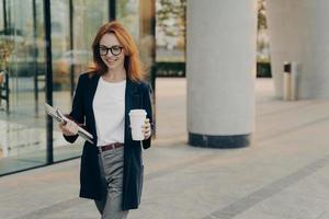 positieve roodharige jonge Europese vrouw in elegante kleding houdt notebookpen en laptop vast foto