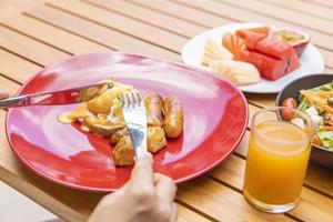 vrouwenhand die een vork houdt en ontbijt eet. Egg Benedict, fruit zoals watermeloen, papaya, meloen, passievrucht, sinaasappelsap en koffie. op een grijze placemat gelegd foto