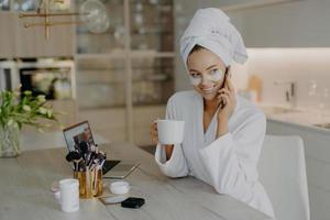 gelukkige jonge europese vrouw ondergaat schoonheidsprocedures na ochtenddouche draagt hydrogel hydraterende pleisters gekleed in witte badjas drinkt aromatische koffie praat via mobiele telefoon poseert binnen foto