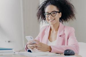 positieve vrouw met krullend haar draagt een bril, formele kleding, houdt moderne mobiele telefoon vast, zoekt informatie over zakelijk nieuws, zit aan bureaupapier, drinkt koffie, heeft een vrolijke uitdrukking, glimlacht foto