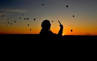 silhouet van slimme telefoon in handen van vrouw die foto's maakt van een prachtig landschap en ballonnen in cappadocië. zonsopgangtijd, dromerig reisconcept foto