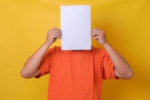 jonge kerel in oranje t-shirt verbergt zijn gezicht onder blanco wit papier, geïsoleerde gele achtergrond. foto