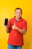 portret van een gelukkige aziatische oude man die een nieuwe smartphone vasthoudt, laat duim omhoog teken zien, stel voor geïsoleerd op gele kleur achtergrond. foto