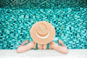 gelukkige vrouw in groene zwembroek en hoed in zwembad bij luxehotel tegen oceaanfront. jonge vrouw geniet in tropische resort. ontspannend, zomer, reizen, vakantie, vakantie en weekend concept foto