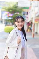 zelfverzekerde jonge aziatische vrouw die een wit overhemd en een schoudertas draagt, glimlacht vrolijk terwijl ze naar haar werk loopt op kantoor in de stad. foto