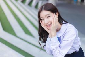 mooie middelbare school Aziatische student meisje in het schooluniform staat en glimlacht gelukkig met beugels op haar tanden terwijl ze haar hand vol vertrouwen op het gezicht legt met het gebouw als achtergrond. foto