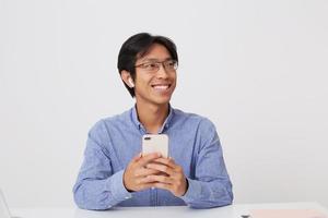 Vrolijke aantrekkelijke Aziatische jonge zakenman in glazen en blauw shirt met behulp van mobiele telefoon en luisteren naar muziek met draadloze koptelefoon op witte achtergrond foto