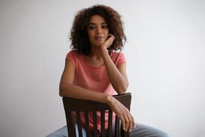 indoor portret van mooie donkere vrouw met bruin krullend haar zittend op een stoel op witte achtergrond, kijkend naar camera met kalm gezicht en lichte glimlach, handpalm onder haar kin houdend foto