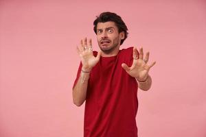 bang bang jonge man met haren in rood t-shirt maken een bang gebaar met zijn handpalmen terwijl hij probeert zichzelf te verdedigen over roze achtergrond foto