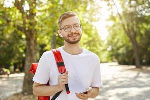 mooie bebaarde man in wit t-shirt met mobiel wegkijkend met zachte glimlach, koptelefoon en bril dragend, staande over de groene bomen foto