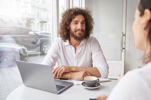 vrolijke jonge man met baard en bruin krullend haar ontmoet vriend in coffeeshop, op afstand werkend met moderne laptop, zittend aan tafel bij raam met gevouwen armen foto