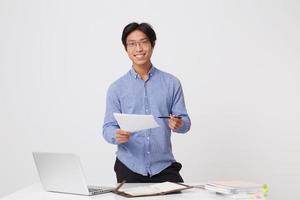 vrolijke aziatische jonge zakenman in glazen die met documenten werkt en camera bekijkt die zich over witte achtergrond bevindt foto