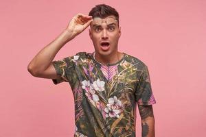 binnenfoto van een verraste mooie jonge kortharige man in een gebloemd t-shirt die over een roze achtergrond staat, een bril op het voorhoofd beweegt met een verbaasde blik foto