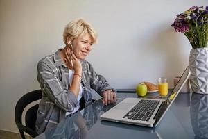 indoor portret van mooie jonge vrouw met blond kort haar, zittend aan tafel, ontbijten met croissants en sap, genieten van video met koptelefoon op haar laptop foto
