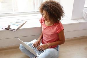 portret van een jonge, mooie gekrulde vrouw met een donkere huid die op de vloer zit met een laptop, de handen op het toetsenbord houdt, zich voor een groot raam poseert, een spijkerbroek en een roze t-shirt draagt foto