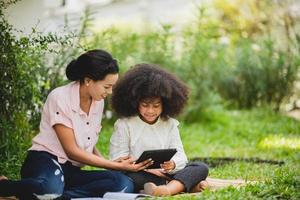 vrolijke jonge mooie moeder zit met zoon met krullend haar in het park en helpt hem tijdens het gebruik van digitale tablet tijdens het studeren foto