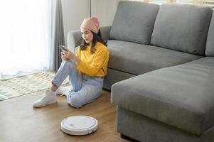 jonge gelukkige vrouw die ontspant en smartphone gebruikt in de woonkamer terwijl robotstofzuiger aan het werk is foto