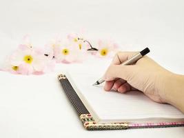 de hand van de dame schrijft dagboek met roze kunstbloem over witte achtergrond foto
