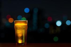 een glas bier met zijn schuim zet op houten tafel van de bar geïsoleerd op een donkere nachtachtergrond met kleurrijke bokehlichten op de bar op het dak. foto