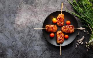 Grill spies melk varkensvlees in een zwarte schotel met tomaten met knoflook en lente-ui op een grijze betonnen ondergrond. Thais straatvoedsel. Bovenaanzicht en kopieer de ruimte foto