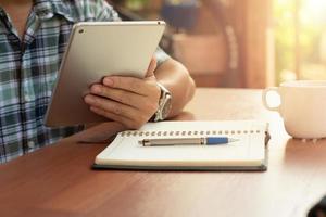 jonge man zit thuis aan tafel, maakt gebruik van digitale tablet op bureau en notebook, kopje coffee.man werken, studeren. foto