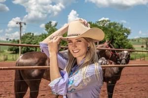 mooie blonde veedrijfster met hoed die zich dichtbij de achtergrond van de paardenranch bevindt foto