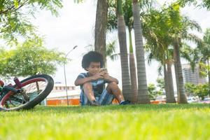 de jonge jongen zit op het gras in de tuin en gebruikt smartphone. een fiets liggend op het gras onder de boom. foto