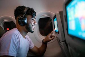 passagier in vliegtuig wat betreft lcd-entertainmentscherm. latijns-amerikaanse man in vliegtuigcabine met behulp van een slim apparaat dat naar muziek luistert op een koptelefoon. foto