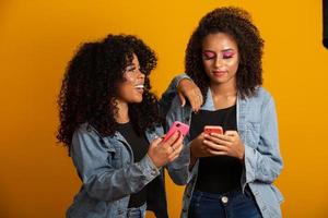 afbeelding van emotionele opgewonden afro jonge vrouwen vrienden geïsoleerd over gele muur achtergrond met behulp van mobiele telefoons. foto