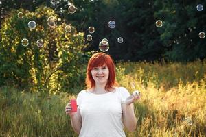 jonge roodharige vrouw zeepbellen blazen. gelukkig meisje in de natuur in de zon. foto