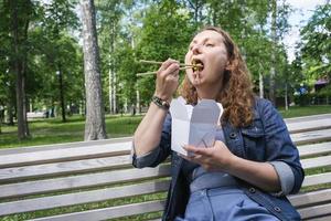 een volwassen vrouw van middelbare leeftijd tijdens een wandeling op een zomerse dag in het park luncht met noedels wok foto