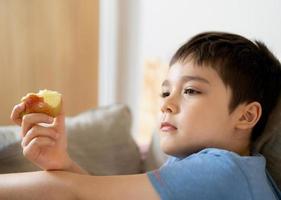 gezond kind dat rode appel eet, schattige jongen die vers fruit eet voor zijn snack terwijl hij tv kijkt in de woonkamer, close-up kind gezicht etend. gezond voedsel voor kinderen concept foto