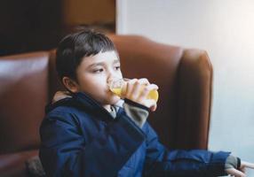 gezonde jongen die vers sinaasappelsap drinkt in het café met fel licht dat uit het raam schijnt, kind zit op de bank in de coffeeshop en kijkt uit met een denkend gezicht terwijl hij sap drinkt na een snack. foto