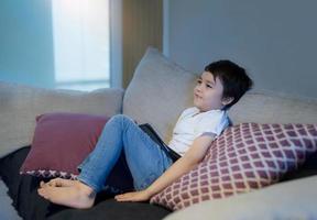 gelukkige jongen met lachend gezicht zittend op de bank kijken naar tekenfilm, schooljongen zit alleen op de bank met tablet en tv kijken, portret kind ontspannen in de woonkamer na terug van school. foto