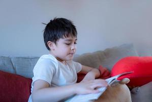 portret gelukkige jonge jongen zittend op de bank een schoolboek lezen, kind geniet van het lezen van verhaal ontspannen thuis, thuisonderwijs concept foto