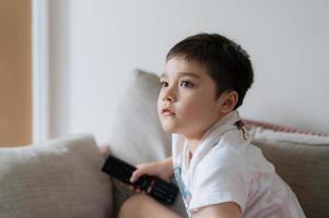 portret jonge jongen die afstandsbediening vasthoudt en omhoog kijkt met een nieuwsgierig gezicht, kind zittend op de bank kijkend naar tekenfilm op tv, kind liggend op de bank ontspannend in de woonkamer na terug van school. foto