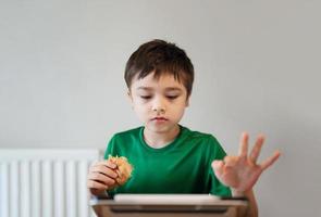 kindjongen die croissant eet en tekenfilm kijkt op tablet, kind aan het ontbijten en playong-spel online op internet met vrienden voordat hij 's ochtends naar school gaat foto